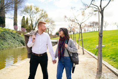Mutlu genç çift el ele tutuşup nehir kenarındaki parkta yürüyor güneşli bir bahar gününün tadını çıkarıyor.