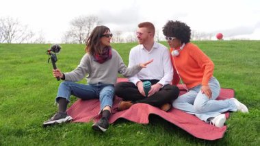 Genç içerik yaratıcıları video blogu çekerken, güneşli gökyüzünün altında çimen battaniyesinde otururken, yaratıcı açık hava yaşam tarzı içeriğini kaydederken iş birliği yapıyorlar.