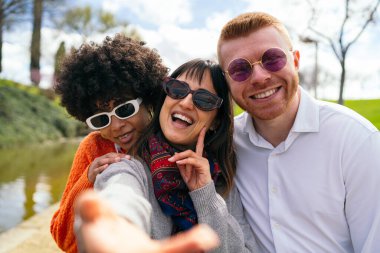 Üç neşeli arkadaş parkta selfie çekiyorlar, güneşli havanın ve güzel arkadaşlığın tadını çıkarıyorlar.