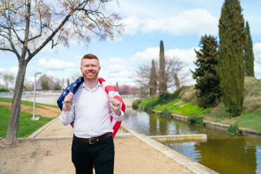 Beyaz tişörtlü ve siyah pantolonlu kızıl saçlı bir adam Amerikan bayrağını omuzlarında taşıyor, gülümsüyor ve parkta yürüyor.