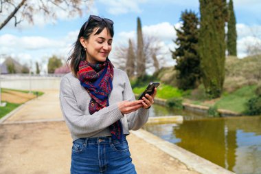 Genç bir kadın, arka planda ağaç ve gölet olan bir parkı ziyaret ederken akıllı telefon uygulaması kullanıyor.
