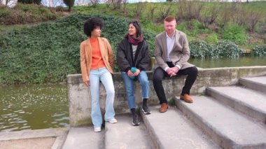 Üç arkadaş, iki kadın ve bir adam, dinlenip göletin kenarında sohbet ediyorlar, güneşli bir günde kahvenin tadını çıkarıyorlar, ağır çekimde yakalanmışlar.