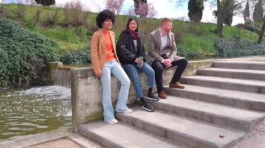 Üç arkadaş, iki kadın ve bir adam, parkta bir göletin kenarında oturmuş, canlı sohbetler yapıyorlar, yavaş çekimde yakalanmışlar.
