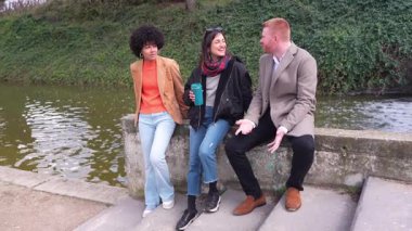 Üç arkadaş, iki kadın ve bir adam, parkta gölün kenarında sohbet ediyorlar, ağır çekimde yakalanmışlar.