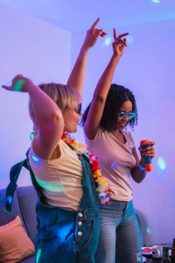 İki çok ırklı bayan arkadaş karaoke yapıyor ve renkli disko ışıklarıyla dans ediyor, eğlenceli bir ev partisinin tadını çıkarıyorlar.