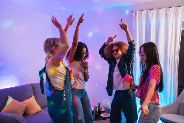 Dört neşeli çoklu etnik arkadaş, renkli disko ışıkları altında şarkı söyleyip dans ederek evde karaoke yapıyorlar.