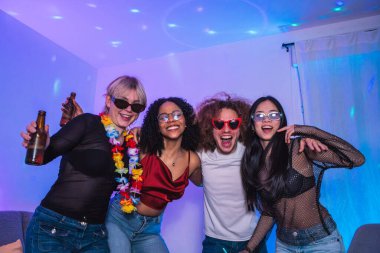 Dört neşeli çoklu etnik arkadaş canlı bir parti atmosferinin keyfini çıkarıyorlar, ellerinde bira şişeleri ve renkli aksesuarları dinamik ışık altında tutuyorlar.