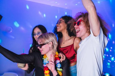 Bir grup neşeli çoklu etnik arkadaş bir ev partisinde selfie çekiyor, renkli disko ışıkları altında müzik ve içkilerin keyfini çıkarıyorlar.