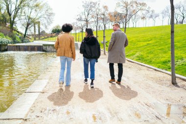Üç arkadaş güneşli bir günde bir göletin yanındaki patika boyunca kameradan uzaklaşıp parkta keyifli bir gezinti yapıyorlar.