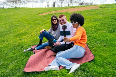 Parktaki bir battaniyenin üzerinde oturan üç içerik yaratıcısı, harici mikrofonlu bir akıllı telefon kullanarak bir video podcast kaydediyor.