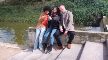 Çok ırklı arkadaşlar gölet kenarında neşeli bir selfie anı yakalıyorlar. Canlı bir dostluk ve gevşek bir bağlantıyla güneşli bir açık hava keyfi sürüyorlar.