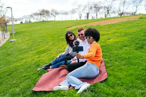 Parkta güneşli bir günün tadını çıkaran üç arkadaş, akıllı telefon ve mikrofonla canlı bir video bloğu çekiyorlar, kahkaha ve yaratıcılığı paylaşıyorlar.