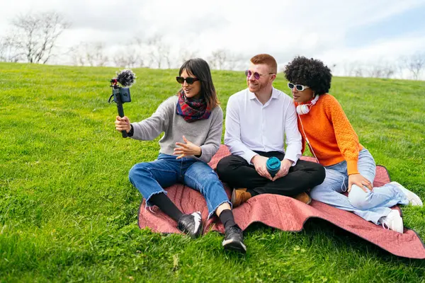 Üç içerik yaratıcısı parkta battaniyenin üzerinde oturmuş akıllı telefon ve harici mikrofon kullanarak video blogu çekiyorlar.