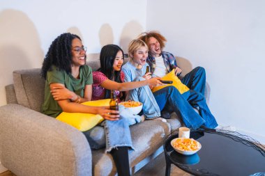 Bir grup neşeli çoklu etnik arkadaş birlikte rahatlatıcı bir akşamın tadını çıkarıyor, televizyon izliyor, bira içiyor ve rahat bir ev ortamında rahat bir kanepede abur cubur yiyor.