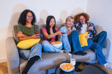 Dört çok ırklı genç arkadaş bir kanepede dinleniyor, televizyon izliyor, sıcak bir ev ortamında içecek ve atıştırmalıkların keyfini çıkarıyorlar.