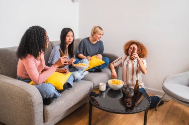 Bir grup neşeli çoklu etnik arkadaş kanepe ve yerde dinleniyor, evde birlikte vakit geçirirken içki ve atıştırmalıkların tadını çıkarıyorlar.