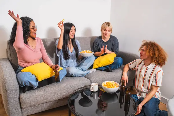 Dört neşeli, etnik çeşitliliğe sahip genç arkadaş bir kanepede dinleniyor, evde birlikte eğlenirken atıştırmalıkların ve içkilerin tadını çıkarıyorlar.