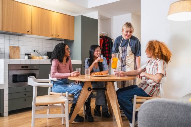 Dört genç etnik çeşitlilikte arkadaş modern bir mutfakta bir masanın etrafında oturmuş kahvaltı ve sohbetin tadını çıkarıyorlar.