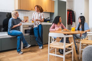 Dört genç etnik çeşitlilikte arkadaş sohbet ediyor ve modern bir mutfakta kahvaltı yapıyorlar, birlikte vakit geçiriyorlar.