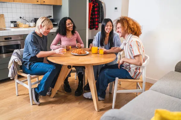 Dört genç çok ırklı arkadaş yuvarlak ahşap bir masada oturmuş modern bir dairede kahvaltı ve sohbetin tadını çıkarıyorlar.