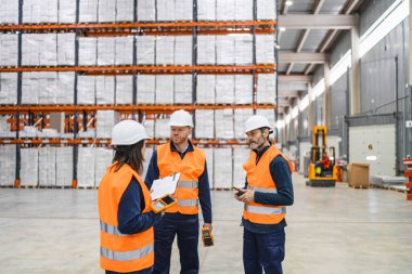 Güvenlik yelekleri ve kask giyen depo çalışanları büyük bir dağıtım merkezinde envanter ve lojistik işlerini yürütmek için barkod tarayıcıları ve panolar kullanıyorlar.