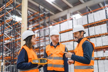 Güvenlik yelekleri ve şapka giyen depo çalışanları lojistiği tartışıyor ve büyük bir depo veya dağıtım merkezinde el bilgisayarı kullanıyorlar.
