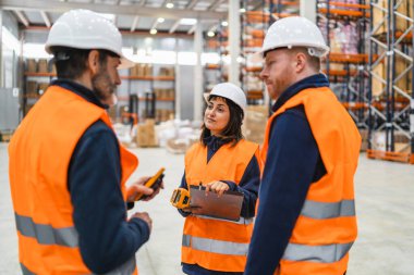 Güvenlik yeleği ve şapka giyen depo çalışanları lojistiği tartışıyor, barkod tarayıcıları ve dosya tutuyorlar.