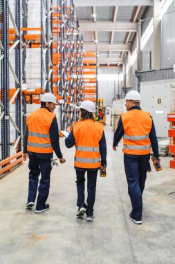 Güvenlik yelekleri ve miğferler giyen depo çalışanları barkod tarayıcısı kullanarak büyük dağıtım merkezinden geçiyor ve lojistik operasyonları tartışıyorlar.
