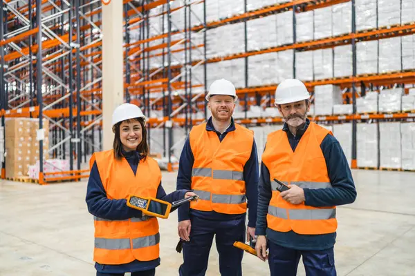 Güvenlik yelekleri ve kasketleri olan depo işçileri büyük bir depoda gülümsüyor ve ellerinde envanter yönetim cihazları tutuyorlar.