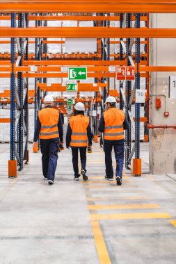 Yüksek görüş açısına sahip yelek ve güvenlik kaskı takan depo işçileri koridorlarda barkod tarayıcılarıyla envanteri kontrol ediyorlar.