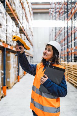 Depo çalışanı barkod okuyucusuyla yüksek rafları tarıyor, büyük bir dağıtım merkezinde envanter ve lojistiği yönetiyor.