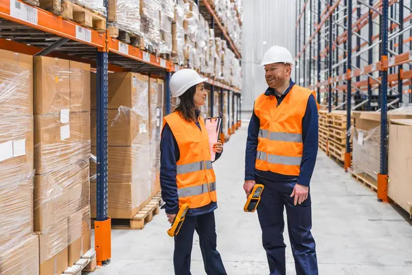 Güvenlik yelekleri ve başlık takan depo çalışanları lojistiği tartışıyor ve envanter kontrolü için barkod tarayıcıları kullanıyorlar.
