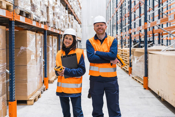 Warehouse workers wearing hardhats and high visibility vests smiling confidently while holding barcode scanners and clipboards in a distribution center
