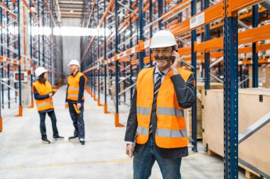 Depo yöneticisi güvenlik yeleği ve kask takıyor. Akıllı telefon kullanıyor ve tablet tutuyor. Lojistik operasyonlarını arka plandaki işçilerle koordine ediyor.