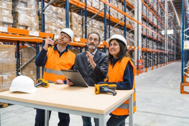 Lojistik merkezinde güvenlik yelekleri ve miğferler giyen depo çalışanlarıyla görüşen bir müdür.