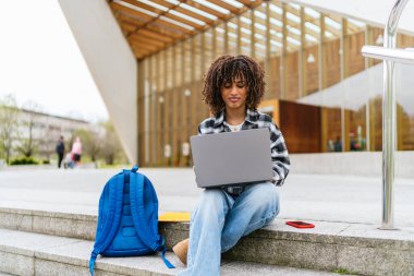 Üniversite basamaklarında otururken dizüstü bilgisayar kullanan Latin kökenli konsantre genç bir üniversite öğrencisi.