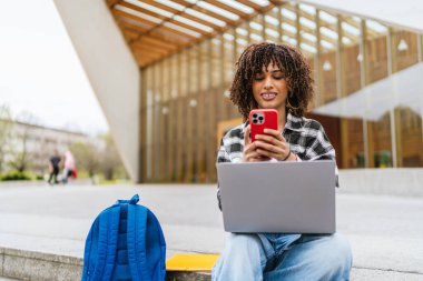 Laptopuyla çalışan Latin kökenli genç bir öğrenci ve üniversite merdivenlerinde otururken akıllı telefonla konuşuyor.