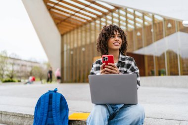 Laptopuyla çalışan Latin kökenli genç bir öğrenci ve üniversite merdivenlerinde otururken akıllı telefonla konuşuyor.