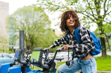Afro saçlı ve sırt çantalı Latin kökenli genç bir öğrenci klasik fotoğraf makinesiyle bisiklet paylaşım servisi kiralıyor.