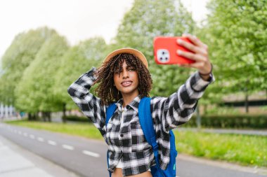 Hasır şapkalı ve sırt çantalı genç bir Latin öğrenci şehir parkında akıllı telefonuyla selfie çekiyor.