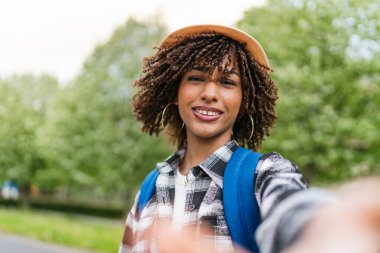 Sırt çantası ve şapkasıyla gülümseyen genç Latin Amerikalı öğrenci, yeşilliklerle dolu canlı bir parkta neşeli bir selfie çekiyor.