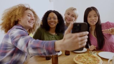 Mutlu çoklu etnik arkadaşlar akıllı telefonlarıyla selfie çekiyorlar. Evde pizza ve içeceklerin tadını çıkarıyorlar.