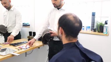 Saç kliniği teknisyenleri kel bir müşteriye farklı peruk ve saç implantları göstererek saç değiştirme seçeneklerini tartışıyor.