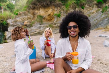 Genç çoklu etnik arkadaşlar plajda dinleniyorlar, meyve suyu içiyorlar ve yaz tatillerinin tadını çıkarıyorlar.