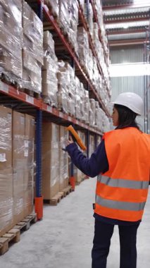 Güvenlik yeleği ve kask giyen deneyimli bir depo çalışanı, düzenli depolama tesisi içindeki paketleri düzenli olarak tarıyor, verimli bir envanter yönetimi ve lojistik iş akışı sağlıyor.