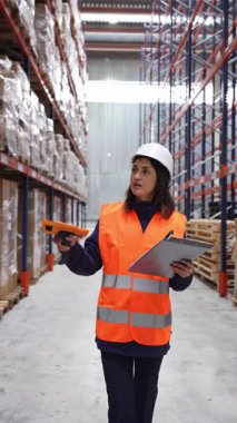 Güvenlik yeleği ve kask giyen bir depo işçisi barkodları tarıyor ve el bilgisayarıyla envanteri kontrol ediyor.