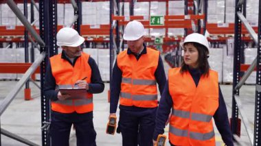 Lojistik ekibi geniş bir depoda iş birliği yapıyor, koridorlarda yürürken envanterleri gözden geçiriyor, güvenlik ekipmanları giyiyor ve el bilgisayarlı izleme cihazları kullanıyor.