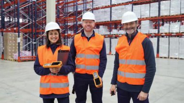 Depo lojistik ekibi, modern depolama tesisinde çalışırken güvenli bir şekilde gülümseyen parlak güvenlik yelekleri ve koruyucu kasklar takıyor.
