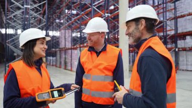 Lojistik işçileri depoda hisse yönetimini tartışıyor, el bilgisayarı kullanıyor ve gülümsüyor.