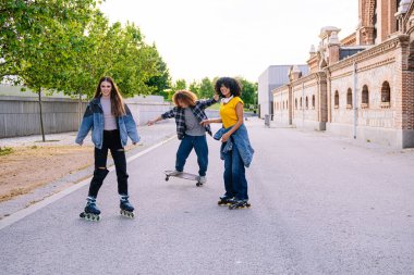 Üç genç arkadaş, öğleden sonra şehir parkında kaykay ve kaykay yaparken eğleniyorlar.